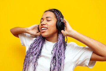 cheerful african american woman with colored dreadlocks listens to music on headphones on yellow...