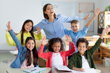 Excited diverse schoolchildren raising arms and smiling, sitting in classroom with their happy young female teacher, back to school concept