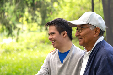 A man and a young man are sitting in a park. The young man is wearing a blue shirt and a gray sweater. Both are smiling and laughing.