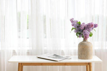Vase with lilac flowers on table near light curtain in living room