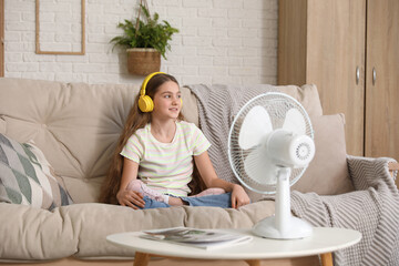 Young girl sitting on sofa near electric fan and listening music in living room