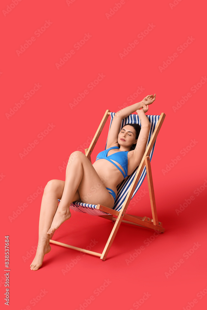 Wall mural Beautiful young woman in swimsuit resting on deckchair against red background