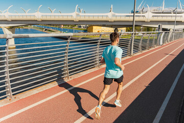 Obraz premium A man jogs on a red pathway along a waterfront, with modern bridges and urban scenery in the background.