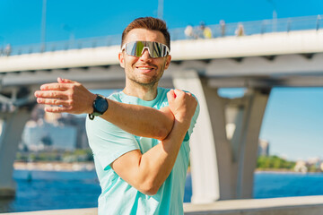 A man stretches his arm while smiling, wearing sunglasses, with a bridge and waterfront in the...