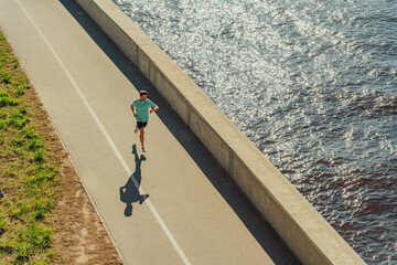 A man jogs along a sunlit waterfront path, casting a long shadow, beside the sparkling water and grassy area.