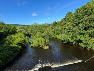 Obraz premium The River Calder flows through a lush landscape, under a blue sky, as gentle ripples on the waters surface, and a small waterfall, create a tranquil scene in, Elland, Yorkshire, UK