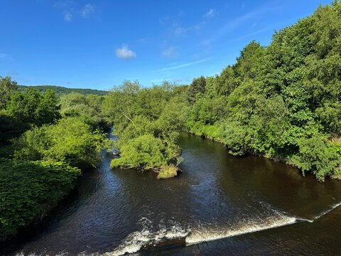 The River Calder flows through a lush landscape, under a blue sky, as gentle ripples on the waters surface, and a small waterfall, create a tranquil scene in, Elland, Yorkshire, UK