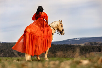 A woman in a orange dress riding on a palomino horse in front of a rural landscape