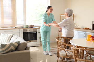 Physical therapist helping senior woman with stick to walk in kitchen