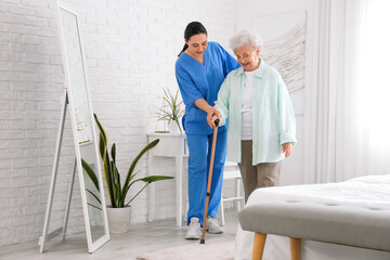 Physical therapist helping senior woman with stick to walk in bedroom