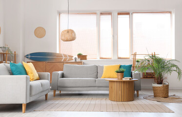 Stylish interior of living room with grey sofa, coffee table, shelving unit and surfboard