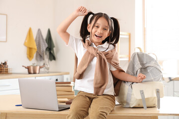 Happy little Asian girl with laptop and backpack sitting on table in kitchen