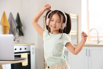 Little Asian girl with headphones and laptop dancing in kitchen