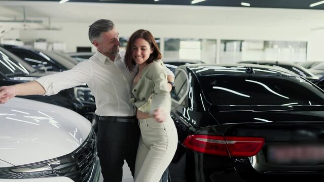 Happy brunette girl in white pants jumps into the arms of her happy middle-aged man in a white shirt after buying their new car in a modern car dealership