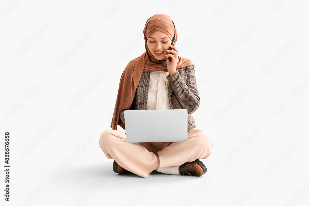 Wall mural Young Muslim businesswoman with laptop talking by mobile phone on light background