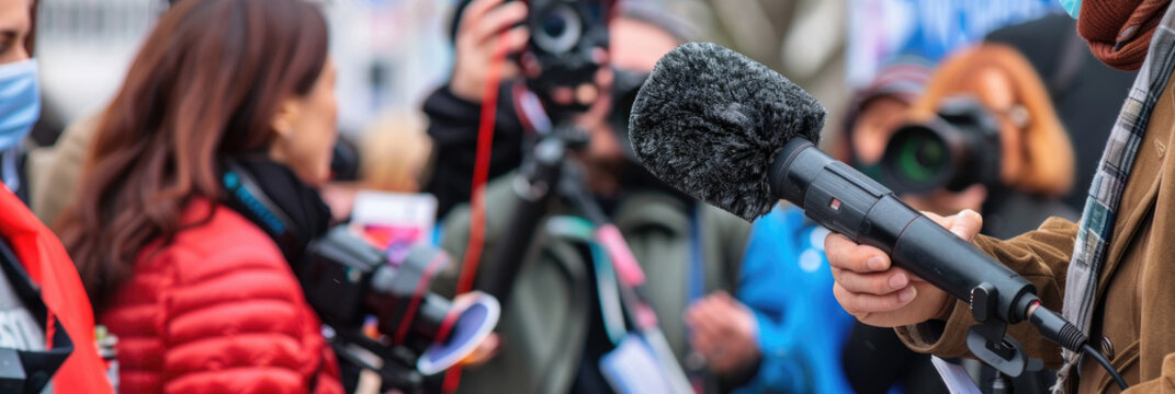 A close-up image of a journalist holding a microphone while interviewing someone at an outdoor event, surrounded by other journalists and members of the public