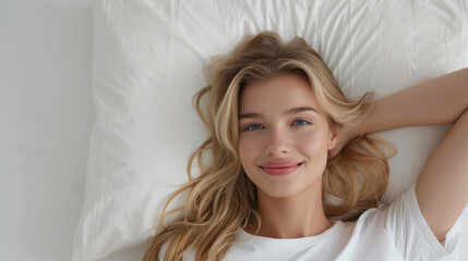 Fototapeta premium Beautiful smiling young blonde female model with long hair and perfect smooth skin relaxing lying on soft white bed looking up