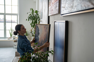 Thoughtful female artist holding painting in frame, hanging on wall. Serious girl creating colorful composition of paintings at apartments, making cozy creative atmosphere in home art studio workshop. © DimaBerlin