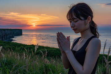 Japanese woman in a serene prayer pose on a grassy hill at dusk. She is 24 years old and has a very beautiful face, in the background is a calm sea