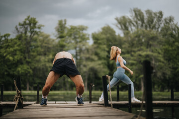 Athletic couple practicing outdoor fitness exercises on a wooden dock surrounded by lush greenery...
