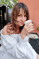 a beautiful woman with dark hair, wearing a long white dress, drinks coffee and laughs cheerfully...
