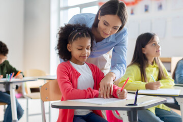 Teacher helping black girl in classroom, black schoolgirl writing in notebook, sitting at desk with...