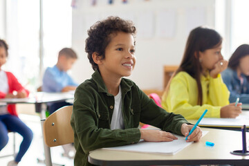 Latin guy writes in exercise notebook and listening teacher, taking notes or writing test. Group of children working diligently and learning