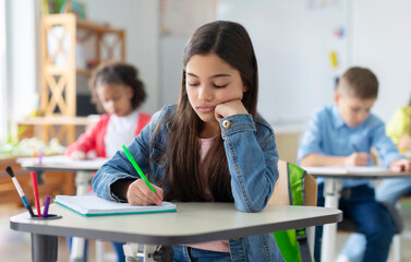 Tired and bored schoolgirl sitting at desk in classroom at school, writing in her notebook and thinking, resting head on hand