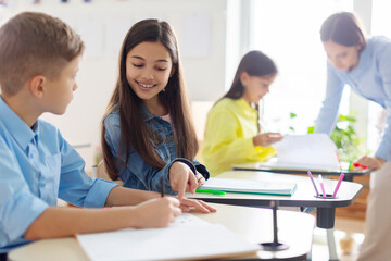 Smart European schoolgirl and boy sitting together at desk in classroom during lesson in private...