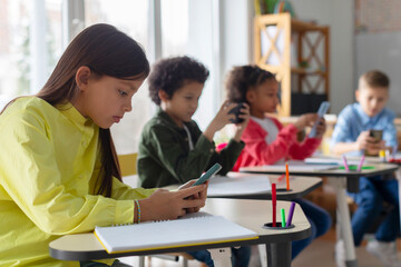 Diverse multiethnic kids students having break and using smartphones, sitting at desks in...