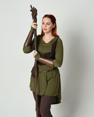  portrait of beautiful red haired female model, wearing green medieval fantasy costume with brown tunic armour armour. Standing pose with wood wizard staff weapon, isolated white studio background.