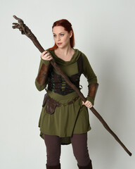  portrait of beautiful red haired female model, wearing green medieval fantasy costume with brown tunic armour armour. Standing pose with wood wizard staff weapon, isolated white studio background.