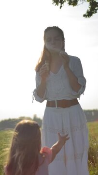 Mom in a white dress blows soap bubbles for her little daughter. They rest in the fresh air, far away from the city. Slow motion handheld medium shot at the sunset. Vertical shot.
