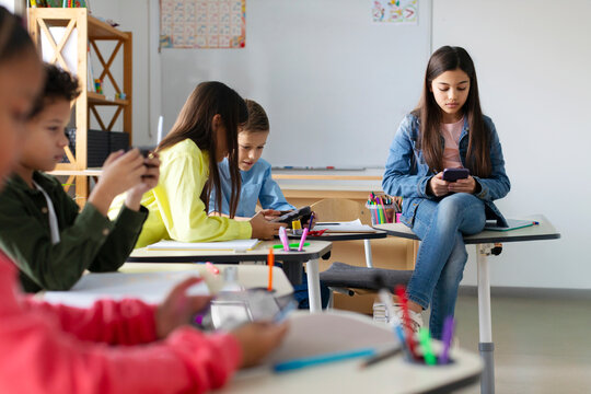 Focused schoolchildren using cellphones while sitting at desks in school classroom interior during break, copy space