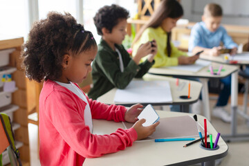 Diverse pupils schoolkids using smartphones in classroom, having break after lessons, children...