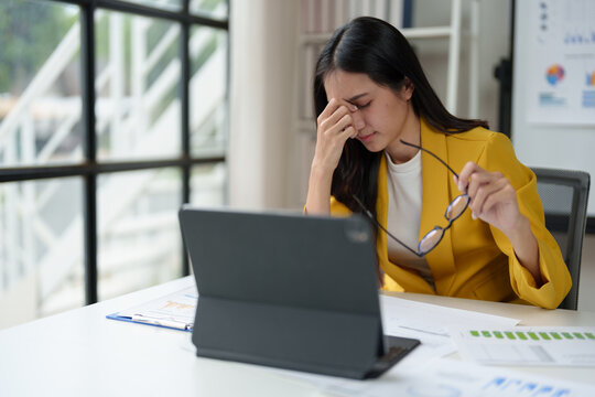Young Asian woman holding glasses has stress, eye strain, headache and fatigue from sitting at a desk for long periods of time with laptop, tablet and paperwork. Office syndrome concept.