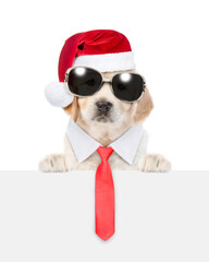 Funny Golden retriever puppy wearing sunglasses, necktie and santa hat looks above empty white banner. isolated on white background