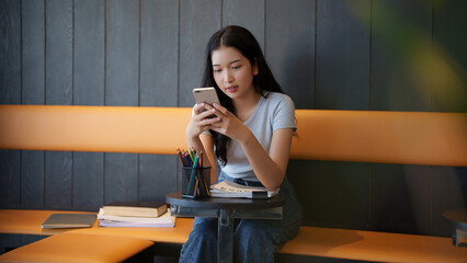 Asian teenage student woman using smartphone to chatting with friends and doing assignment homework after studying and reading information in paperwork to learning about education knowledge in cafe