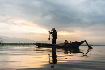 Asian fisherman on wooden boat casting a net for catching freshwater fish in nature river in the early morning before sunrise