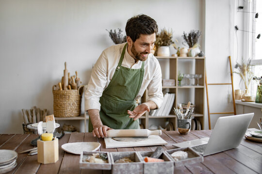 Man rolls clay with rolling pin in a bright and cozy pottery studio. He is concentrated on his work, surrounded by various pottery tools and equipment, creating a relaxed and creative atmosphere.