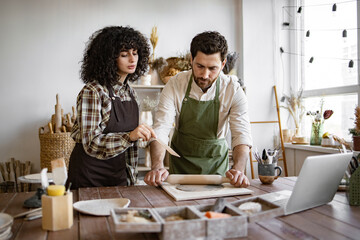 Man rolling clay with rolling pin while woman next to him holds stack in hands, instructing him in pottery studio. Studio filled with various pottery tools and materials, creating creative atmosphere.