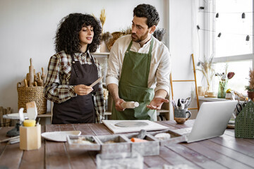 Man rolls clay with rolling pin while woman stands next to him holding stack in hands. Pottery studio with various tools and materials around them, creating handmade ceramic art together.