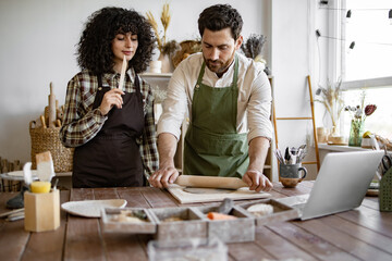 Man rolls clay with rolling pin while woman stands next to him holding stack in hands. Pottery studio with various tools and materials around them, creating handmade ceramic art together.
