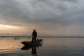 Asian fisherman on wooden boat casting a net for catching freshwater fish in nature river in the early morning before sunrise