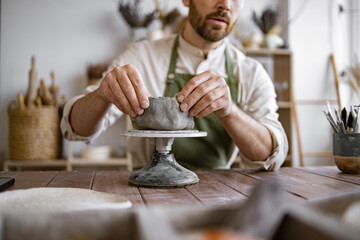 Close up of hands of male artisan wearing an apron works with clay on potter wheel in ceramics workshop. Workspace filled with various pottery tools and materials.