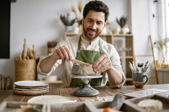 Smiling man working with clay in cozy home pottery studio. Bearded male seated at wooden table with various tools, materials, and laptop in bright well-decorated room.