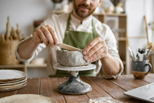 Smiling man working with clay in cozy home pottery studio. Bearded male seated at wooden table with various tools, materials, and laptop in bright well-decorated room.