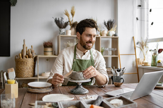 Bearded male seated at wooden table with various tools, materials, and laptop in bright well-decorated room. Smiling man working with clay in cozy home pottery studio.