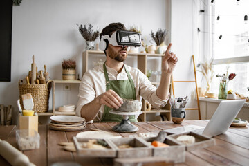Sculptor in pottery studio uses virtual reality glasses to sculpt vase from clay. Workspace filled...