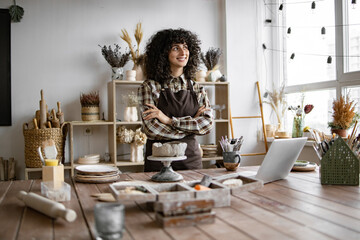 Confident Caucasian female artisan in cozy workspace surrounded by crafting materials, laptop, and decorations, embodying creativity and entrepreneurship.